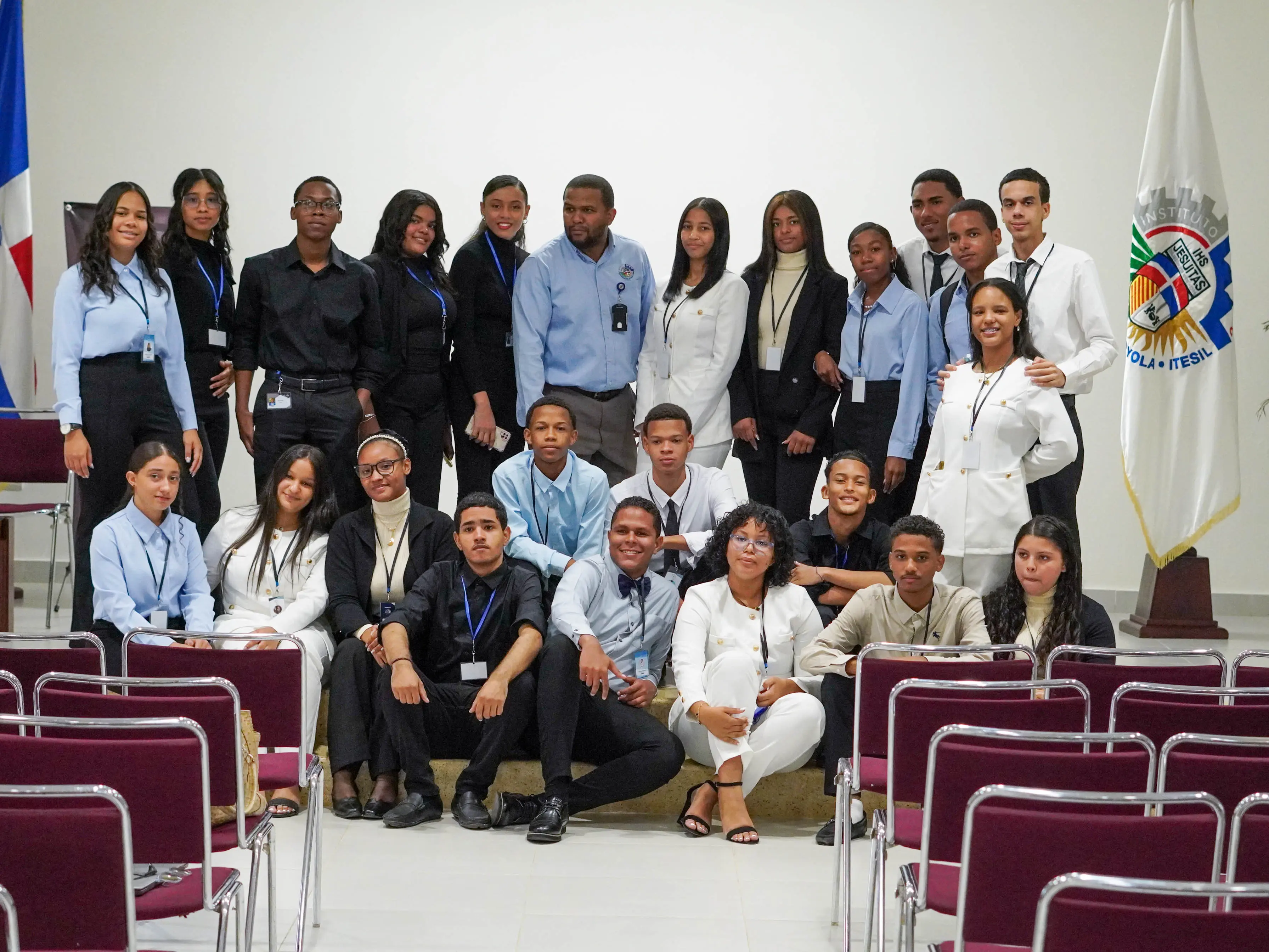 Grupo de estudiantes y docentes del Instituto Tecnológico San Ignacio de Loyola posando juntos en un salón institucional, vestidos formalmente y con gafetes de identificación. Al fondo, se observan las banderas de la República Dominicana y del ITESIL.